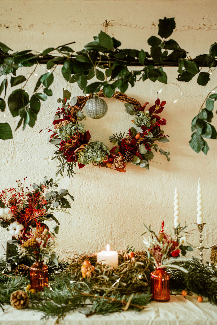 A large dried flower wreath in Christmas colours of red and green hangs on a cream coloured wall behind a table. The table has dried flower arrangements and pine and on it. There are also candles and Christmas tree decorations.