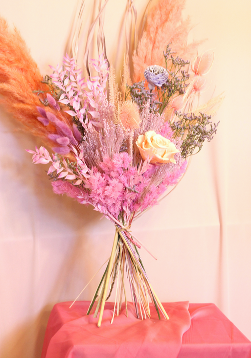 Dried flower bouquet in soft lilac, peach and pink tones. Bouquet is unwrapped and standing against a plain backdrop. 