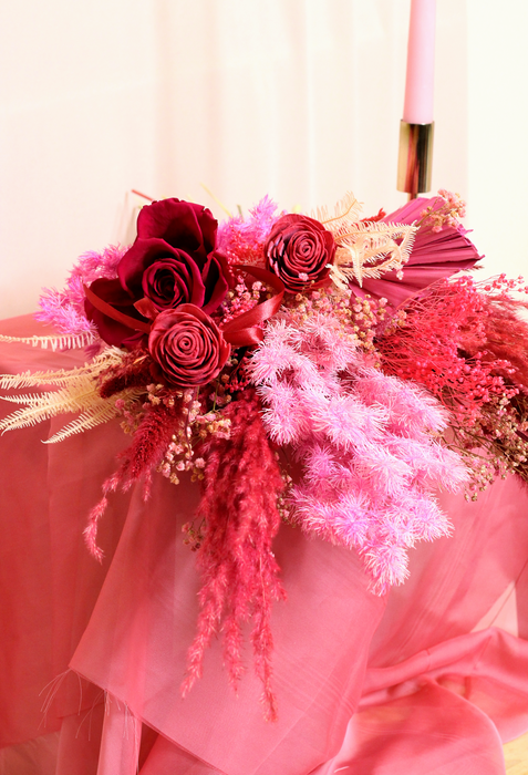 Vibrant dried flower bouquet with dried and preserved flowers. The photo shows a bouquet in deep red and pink tones laying on a surface covered by a pink fabric and set against plain backdrop.