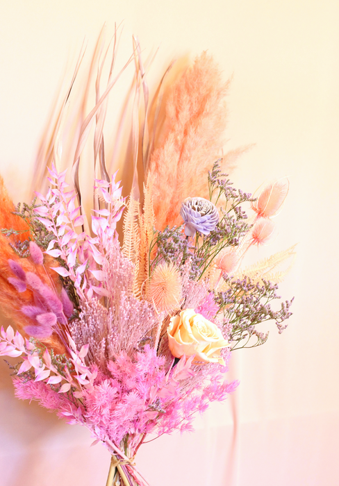 Close up photo of dried flower bouquet in soft pastel tones of lilac, peach and pink. Bouquet is standing against plain backdrop.