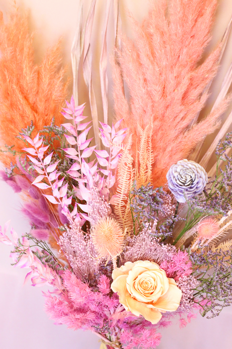Close up photo of dried flower bouquet in soft pastel tones. The photo shows a peach rose on focus among other dried flowers.  Bouquet is standing against plain backdrop.