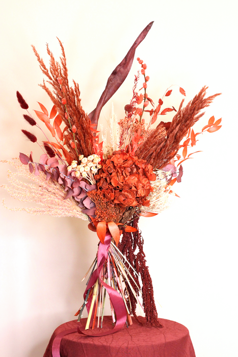 Dried flower bouquet in deep red tones and a touch of soft peach tones. Bouquet is standing against a plain backdrop. 
