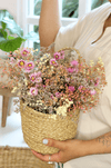 a woman holding a woven jute basket filled with dried flowers (pink daisies and baby's breath)