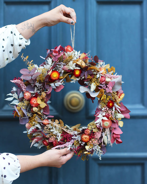 A wreath made of dried flowers is held up by a woman's hands against a background of a blue door