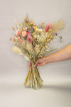 Hand holding dried flower bouquet in natural, pink and green shades, containing helichrysum, thistles, eucalyptus and feathers