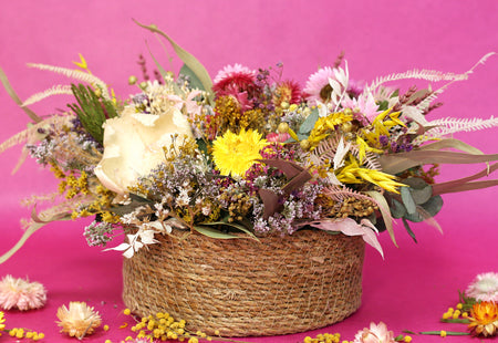 low and round wooven basket filled with colorful dried flowers photographed against pink background