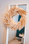 A dried flower pampas wreath in warm neutral tones hangs on a mirror in a white room