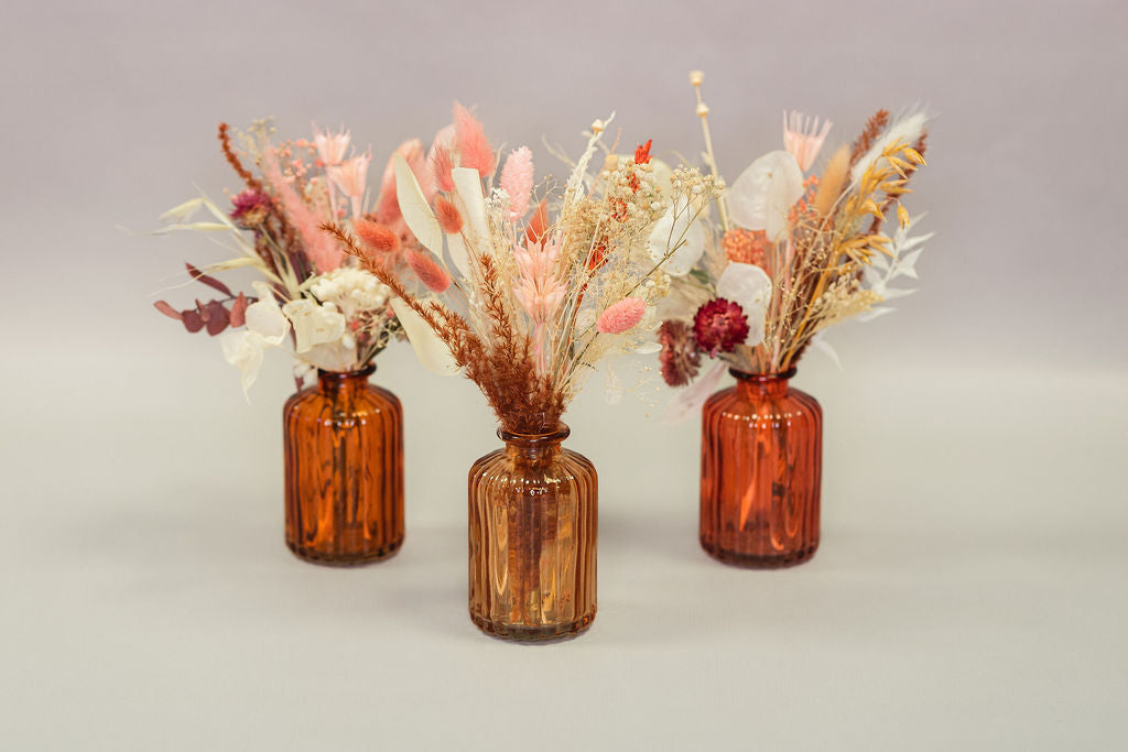 Set of three coloured glass jars in orange tones filled with dried flowers and grasses. 