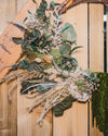 A dried flower wreath made up of eucalyptus, white ruscus, amaranthus and other dried flowers on a twisted wooden wreath.
