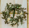 White and green Christmas dried flower wreath featuring eucalyptus, helichrysum, broom bloom and a velvet green ribbon. The wreath hangs on a bamboo frame against a white background.