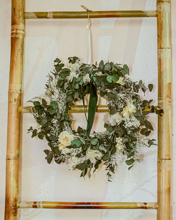 White and green Christmas dried flower wreath featuring eucalyptus, helichrysum, broom bloom and a velvet green ribbon. The wreath hangs on a bamboo frame against a white background.