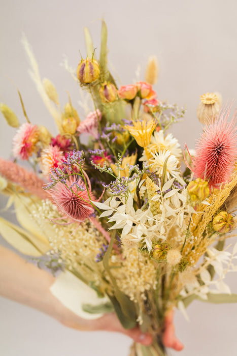 Hand holding dried flower bouquet in natural, green and pink shades, including pink thistles